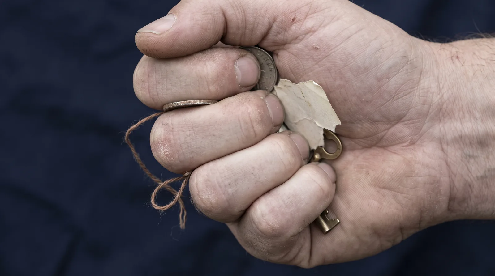 Un puño cerrado con fuerza sosteniendo varios objetos distintos a la vez: una moneda, un papel roto, una llave, un hilo